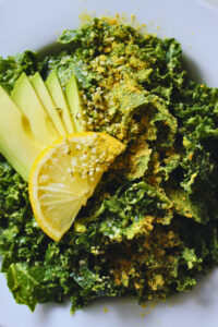 overhead image of kale avocado salad topped with lemon slice, fresh avocado, and a sprinkle of nutritional yeast and hemp seeds